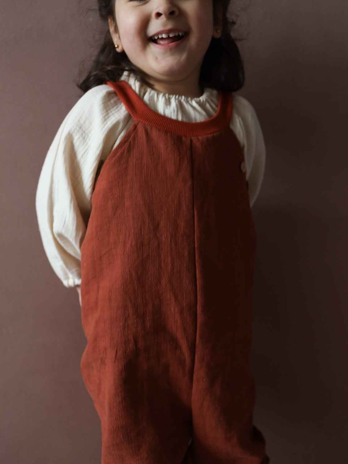 Schlupflatzhose Leinen - Ziegel: Ein Kleinkind trägt locker sitzende, ärmellose Latzhose aus Leinen. Die Hose bietet Bewegungsfreiheit und Komfort, ideal für aktive Kinder im Frühling und Sommer.