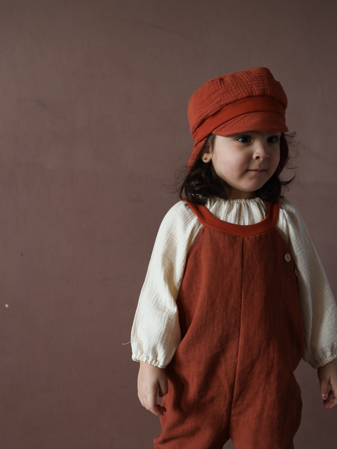 Ein Kleinkind trägt die Schlupflatzhose Leinen - Ziegel, kombiniert mit einer cremefarbenen Bluse und einer passenden Mütze. Die rustrote Hose ist locker geschnitten, ohne Knöpfe oder Reißverschlüsse.