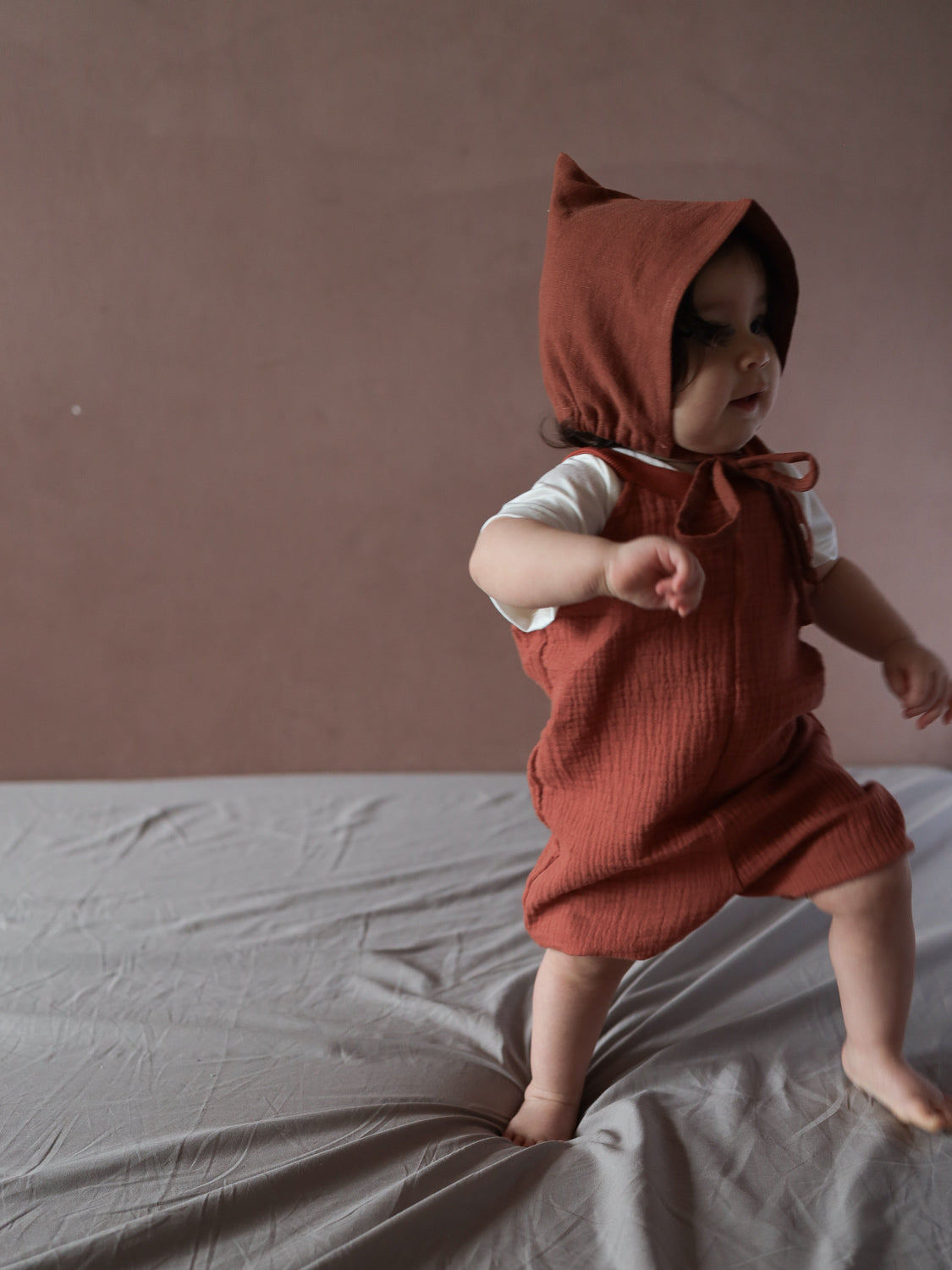 Kleinkind trägt Kurze Schlupflatzhose Bio-Musselin - Terrakotta über weißem T-Shirt, steht barfuß auf einem grauen Laken. Der bequeme Einteiler mit Kapuze ist handgenäht und ideal für den Sommer.