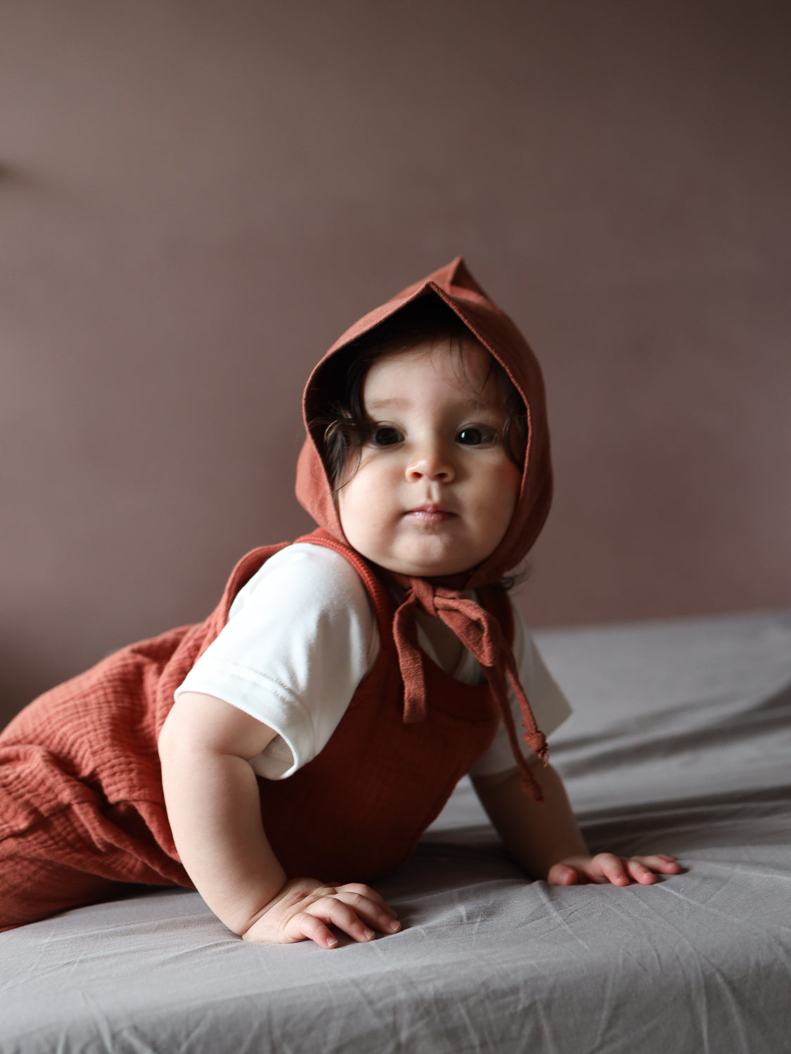Ein Baby trägt die Kurze Schlupflatzhose Bio-Musselin - Terrakotta, einen rustfarbenen Strampler mit passender Kapuze, auf einem weichen Untergrund. Perfekt für den Sommer, handgenäht in Berlin.