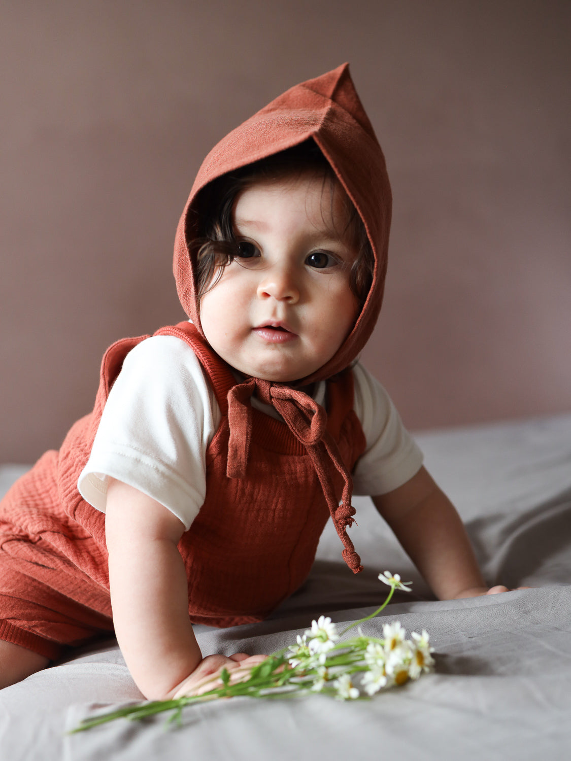 Ein Baby trägt die Kurze Schlupflatzhose Bio-Musselin - Terrakotta, kombiniert mit einem cremefarbenen Shirt und einem Leinenbonnet, während es auf einer weichen Oberfläche neben Gänseblümchen liegt.