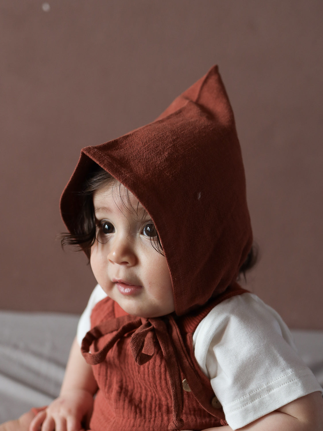 Ein Baby trägt die Kurze Schlupflatzhose Bio-Musselin - Terrakotta, kombiniert mit einem weißen Shirt und passender Mütze, handgenäht in Berlin, ideal für sommerliche Bewegungsfreiheit.