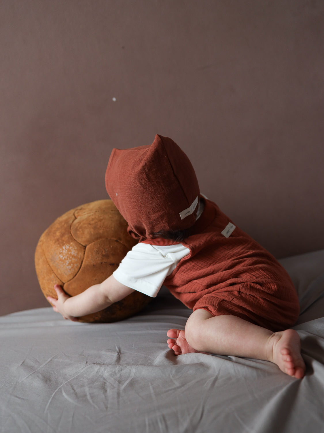 Baby in rustfarbenem Kurze Schlupflatzhose Bio-Musselin - Terrakotta und passender Haube, liegt auf weichem Untergrund, stützt sich auf einen großen, abgenutzten Lederball, ideal zum Spielen und Bewegen.