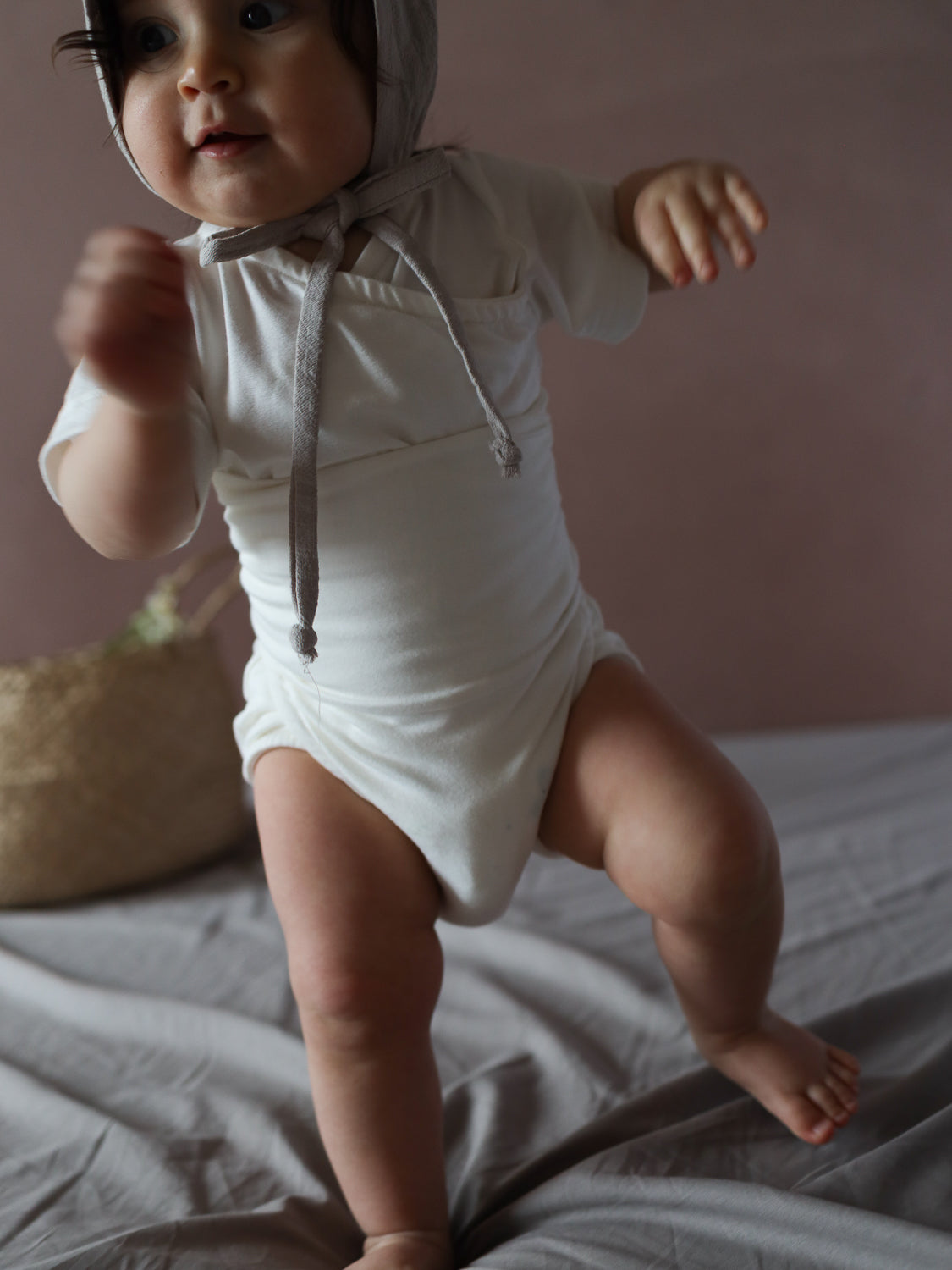 Ein Baby steht auf einem Bett und trägt den Kurzarm Loopbody Bambusviskose - Natur. Der Body ist schlicht mit kurzen Ärmeln, ideal für warme Tage, und schmiegt sich sanft an.