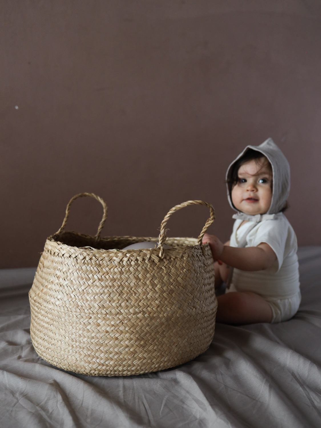 Ein Baby sitzt auf einem Stoffuntergrund, trägt einen Kurzarm Loopbody Bambusviskose - Natur und eine weiche Haube. Vor ihm steht ein großer geflochtener Strohkörbchen.