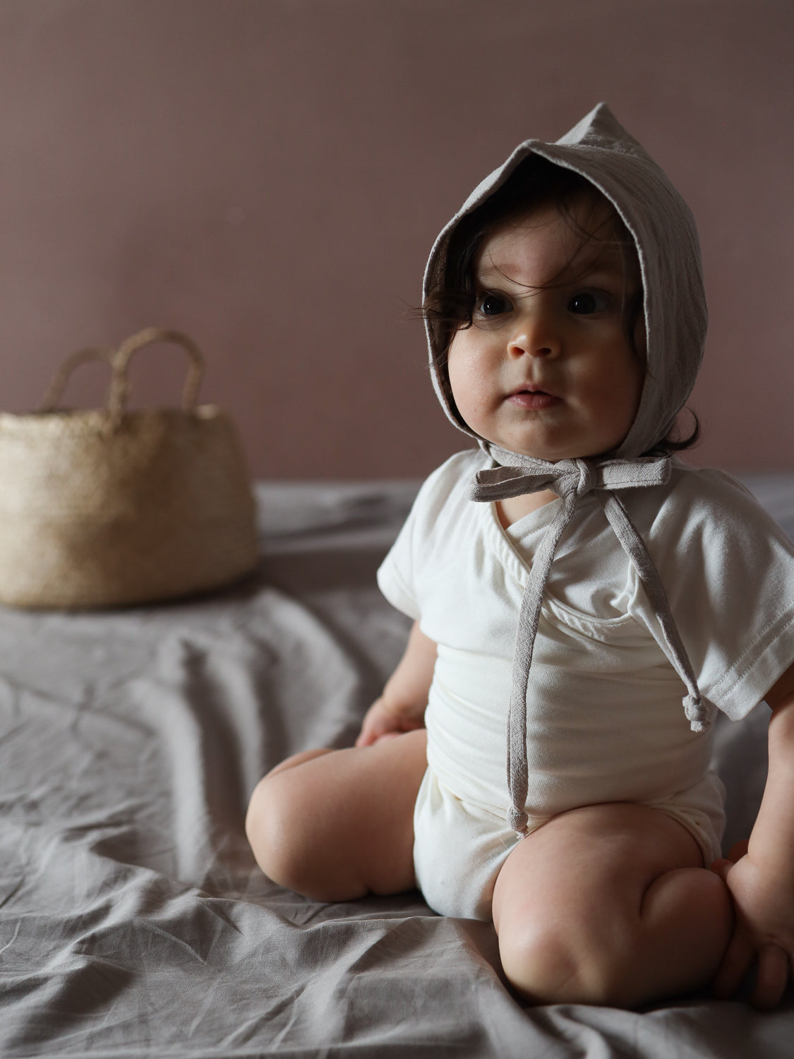 Kleinkind in einem Kurzarm Loopbody Bambusviskose - Natur, sitzt auf einem weichen Bettbezug. Der Body bietet luftige Arme, während der Bauch geschützt bleibt, ideal für warme Sommertage.