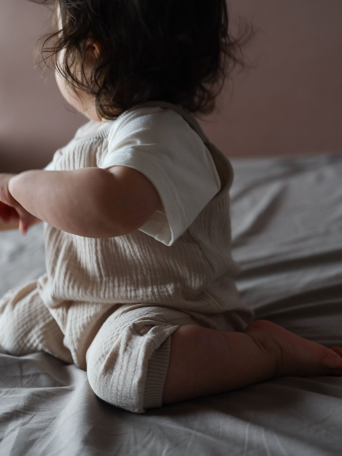 Ein Kleinkind kniet auf einem Bett und trägt die Kurze Schlupflatzhose Musselin - Sand, einen weichen, texturierten Einteiler, der Bewegungsfreiheit bietet und ideal für den Sommer ist.