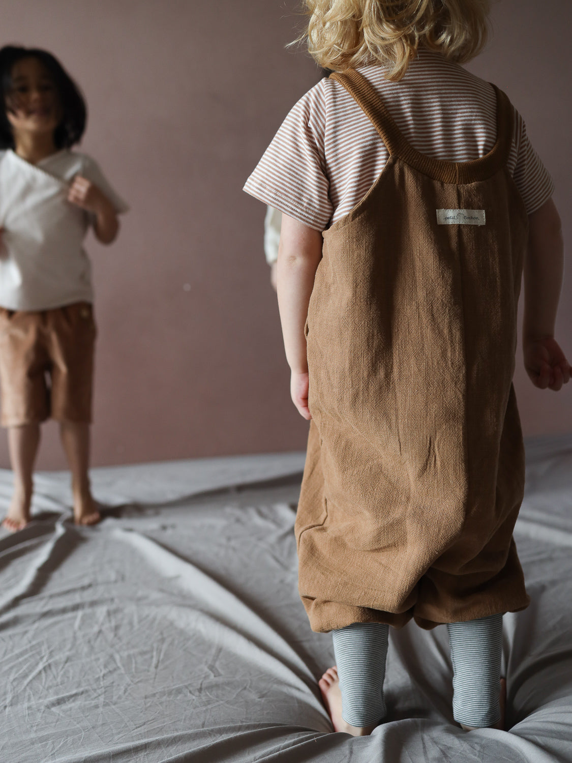 Zwei Kinder stehen barfuß auf einem grauen Stoff. Im Vordergrund trägt ein Kleinkind die Schlupflatzhose Leinen - Weizen über einem gestreiften T-Shirt und Leggings. Minimalistische, neutrale Umgebung.