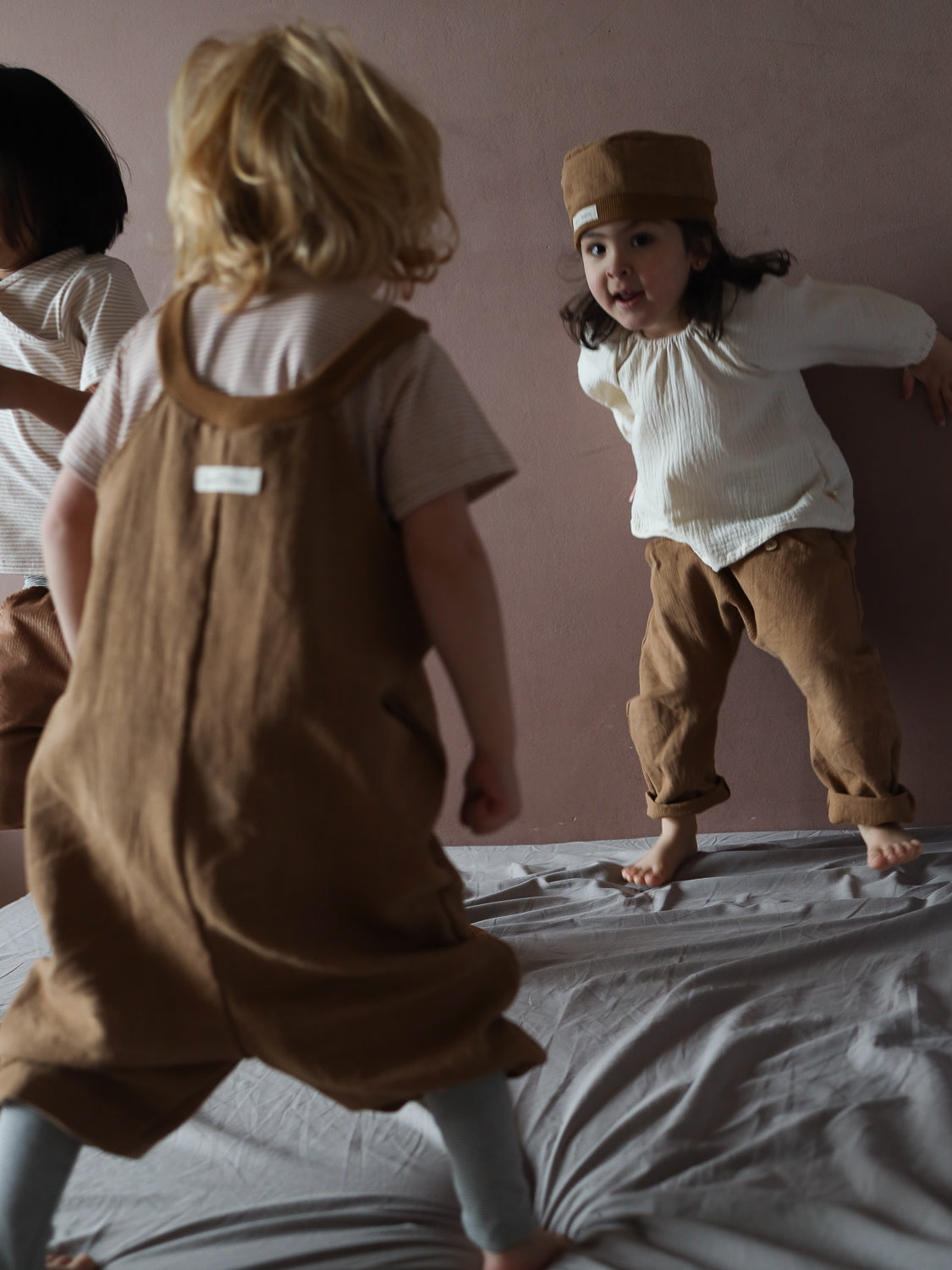 Kleinkinder spielen fröhlich. Eines trägt die Schlupflatzhose Leinen - Weizen, erkennbar an den braunen Overalls aus 100% Leinen. Sie bietet Bewegungsfreiheit, ideal für aktives Spielen.
