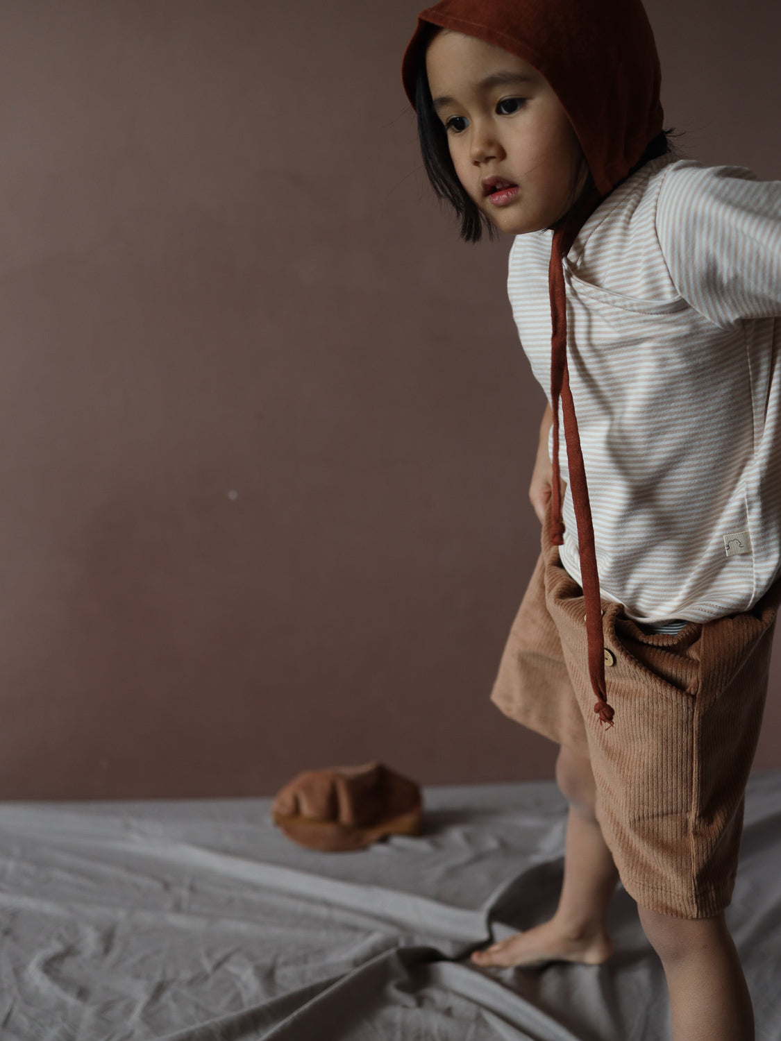 Kleinkind in Kurze Buxe Breitcord - Weizen mit elastischem Bund und camel-brauner Farbe. Trägt gestreiftes Oberteil und rostfarbenes Häubchen, steht auf neutralem Stoff. Minimalistische, erdige Umgebung.