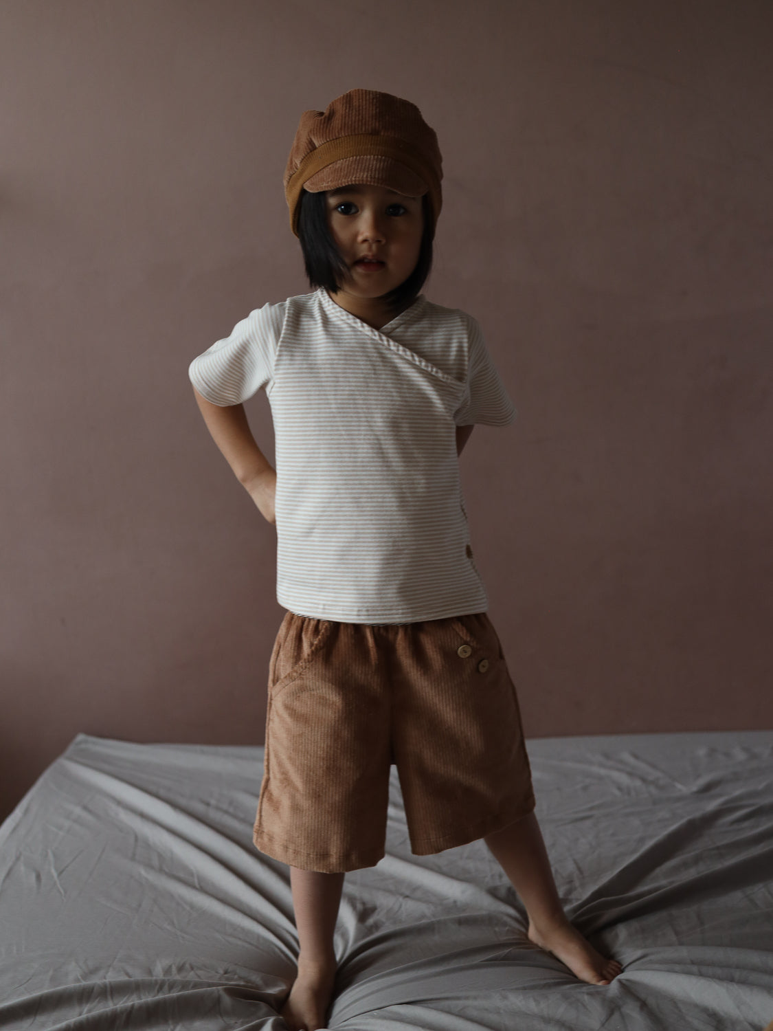 Ein Kind trägt die Kurze Buxe Breitcord - Weizen: braune Cord-Shorts mit Knopfdetails am Hüftbereich, kombiniert mit einem gestreiften Shirt und passender Cap, stehend auf einem Bett.