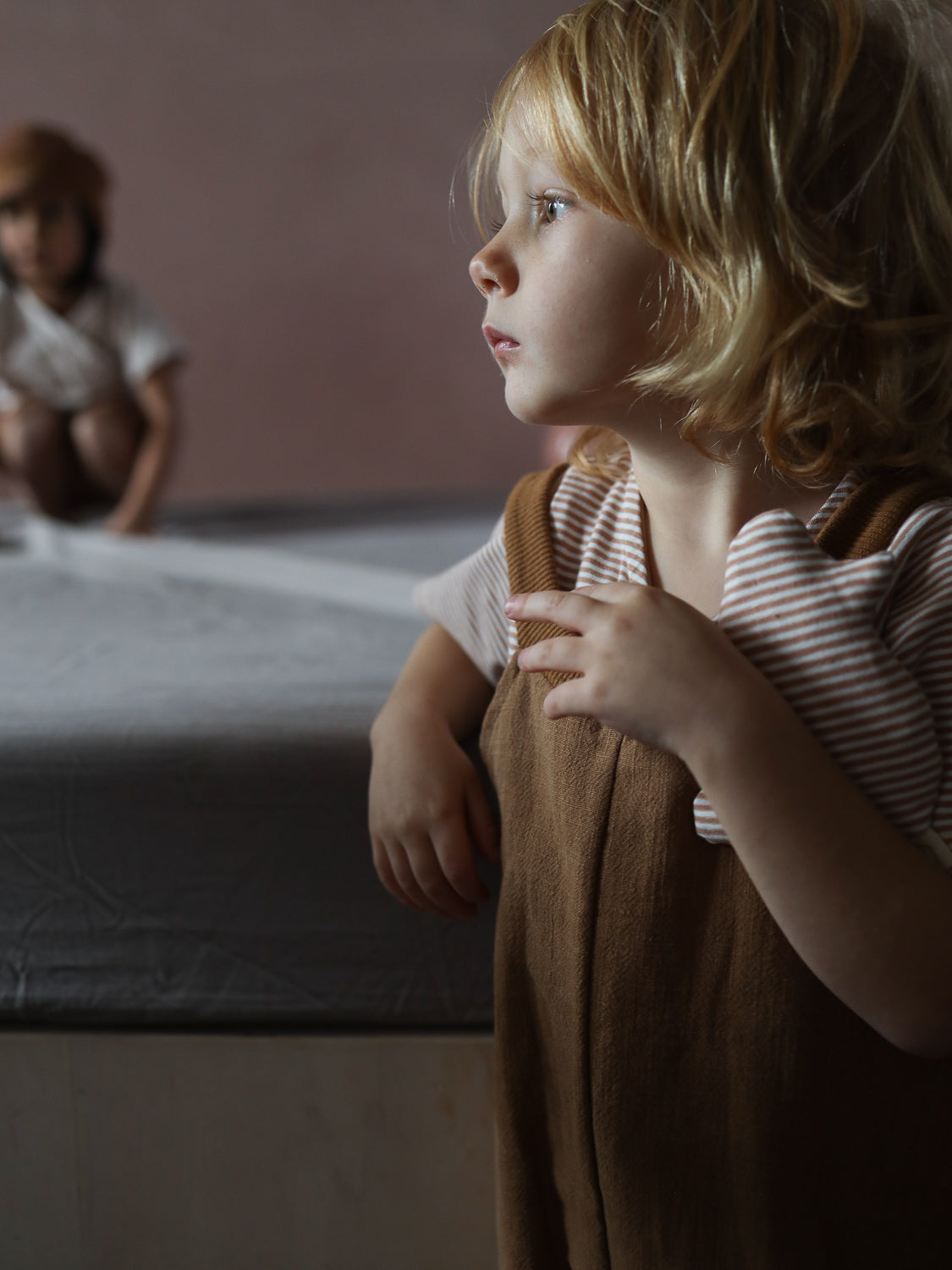 Kleinkind in gestreiftem Kurzes Wickelshirt Baumwolle - Ringel Weizen, unter braunen Latzhosen, steht neben niedrigem Bett. Ein weiteres Kind unscharf im Hintergrund, ruhige, nachdenkliche Atmosphäre.