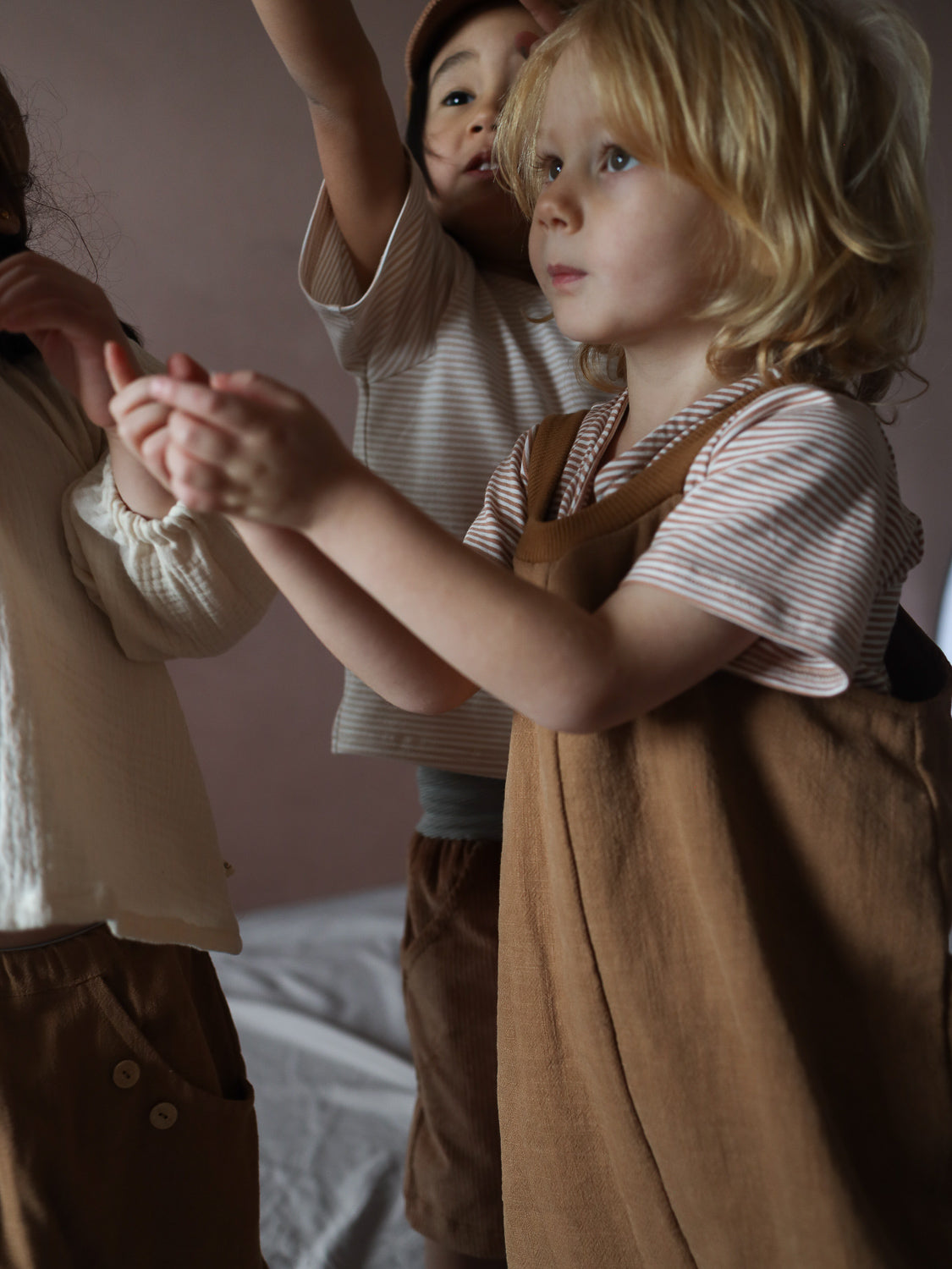 Drei Kinder spielen zusammen, im Vordergrund trägt ein Kind die Schlupflatzhose Leinen - Weizen, kombiniert mit einem beige-weiß gestreiften Shirt, und streckt die Hände aus.