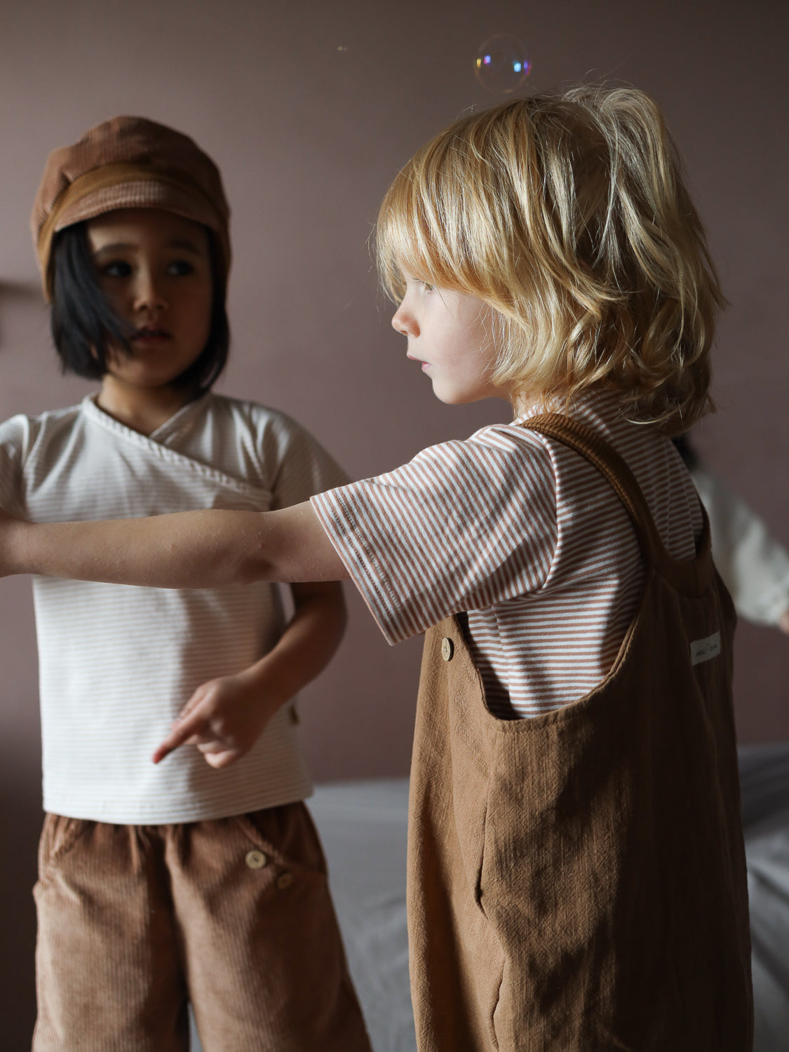 Zwei Kinder spielen drinnen. Das Kind links trägt ein kurzes Wickelshirt Baumwolle - Ringel Weizen, passend zu braunen Cord-Shorts und einer Mütze, während es eine Seifenblase beobachtet.