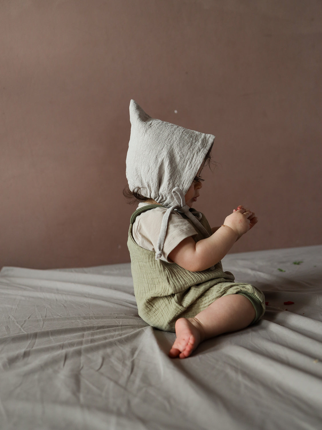 Kleinkind in profil, gekleidet in Kurze Schlupflatzhose Bio-Musselin - Salbei, über beige T-Shirt. Es sitzt barfuß auf weicher Oberfläche und hält einen kleinen Gegenstand, in natürlicher, minimalistischer Kulisse.
