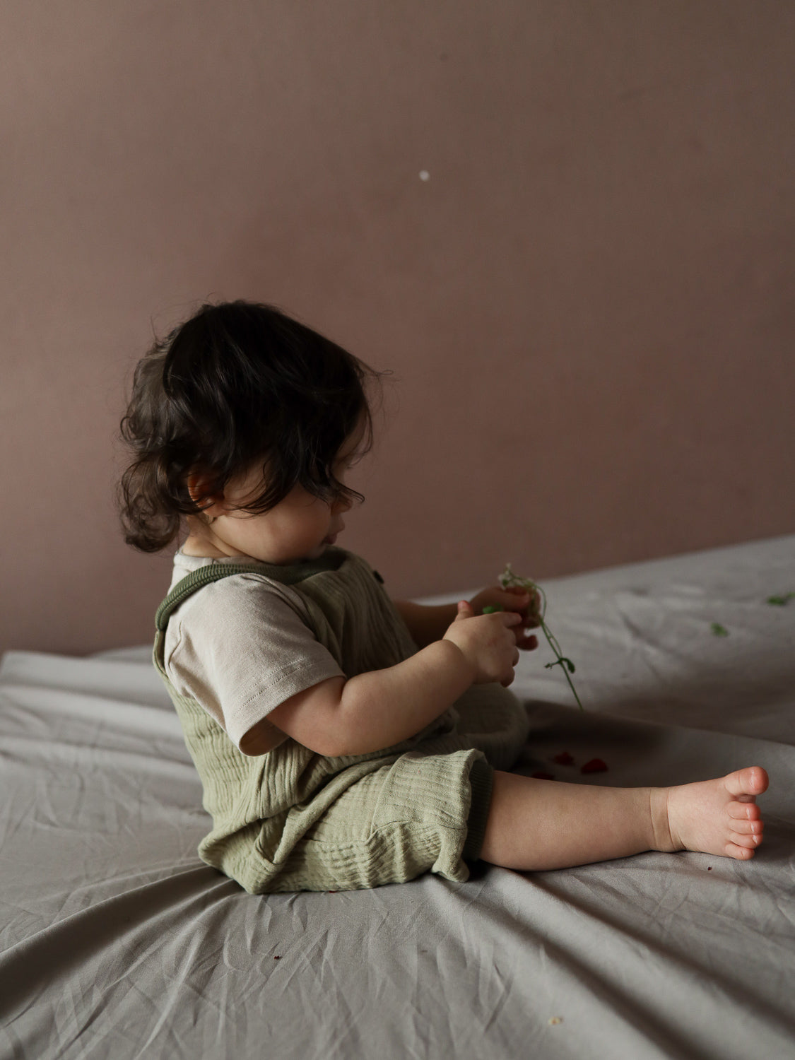 Ein Kleinkind trägt die Kurze Schlupflatzhose Bio-Musselin - Salbei und betrachtet konzentriert einen Pflanzenstängel. Die handgenähte Latzhose bietet Bewegungsfreiheit und ist ideal für den Sommer.