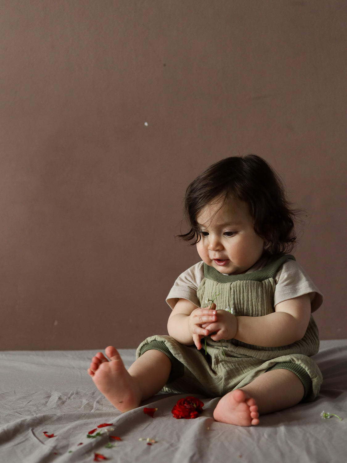 Kleinkind trägt die Kurze Schlupflatzhose Bio-Musselin - Salbei, sitzt barfuß und hält einen Stiel mit weißen Knospen. Die weiche, handgenähte Latzhose bietet Komfort und Bewegungsfreiheit, ideal für den Sommer.