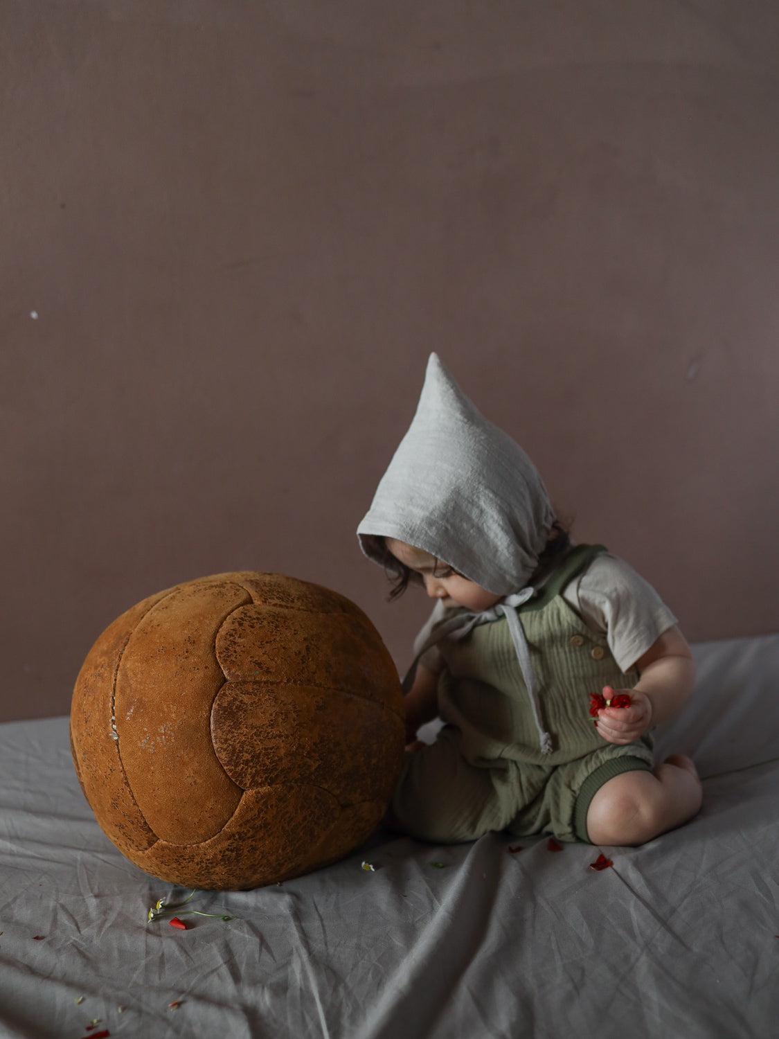 Ein Kleinkind trägt die Kurze Schlupflatzhose Bio-Musselin - Salbei, sitzt auf einem weichen Tuch, hält rote Blütenblätter und befindet sich neben einem großen, lederartigen Ball.