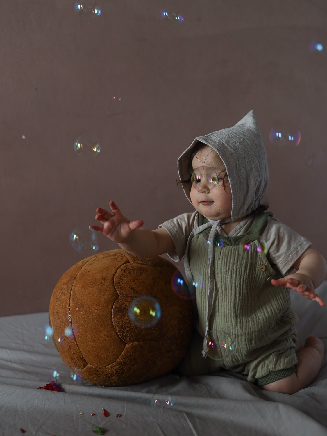 Kleinkind in Kurze Schlupflatzhose Bio-Musselin - Salbei kniet, greift nach Seifenblasen neben vintage Lederball.