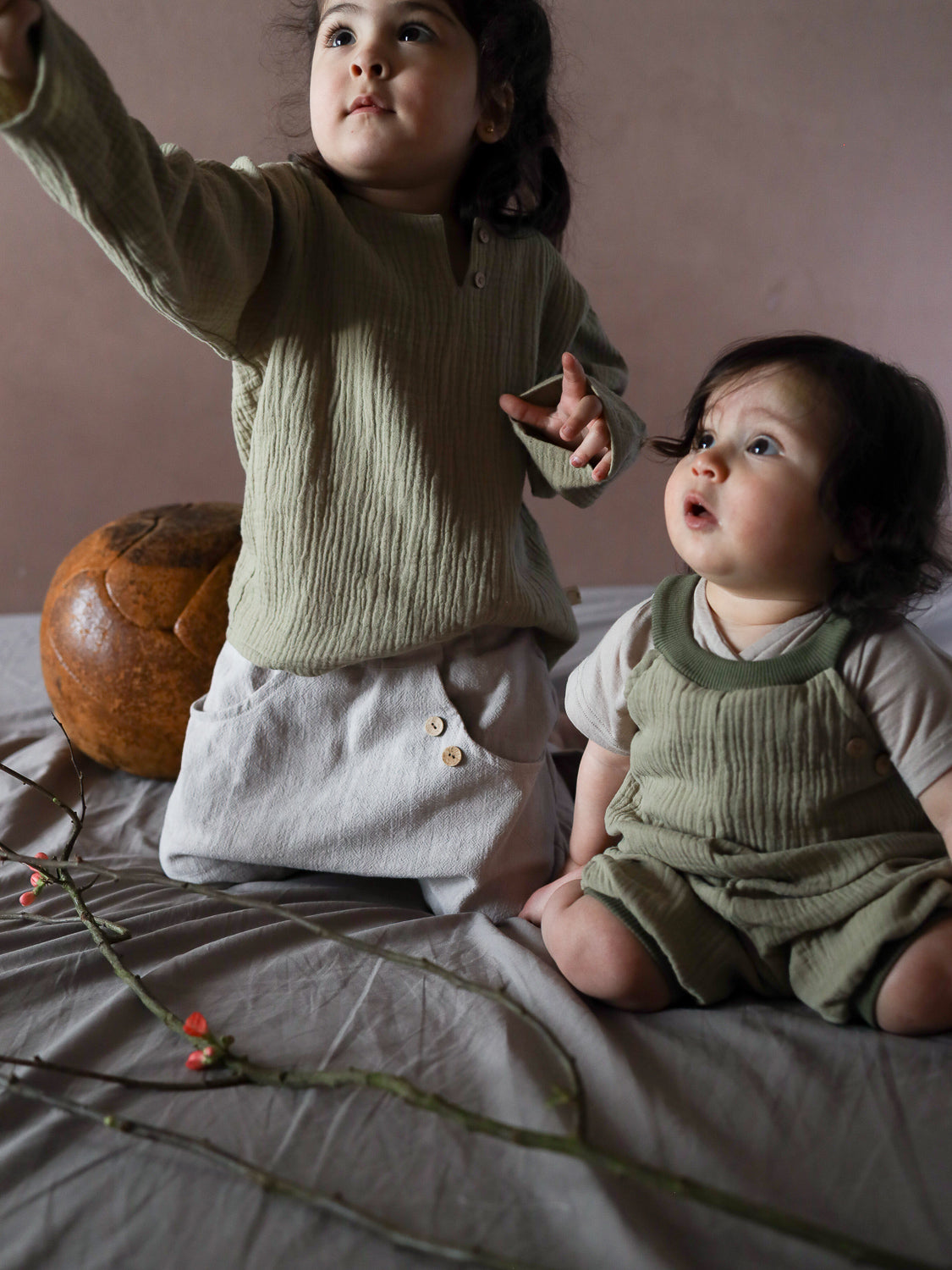 Zwei Kleinkinder in Kurze Schlupflatzhose Bio-Musselin - Salbei aus geknitterter Baumwolle, umgeben von roten Blütenzweigen, mit einem runden Dekorationsobjekt im Hintergrund.