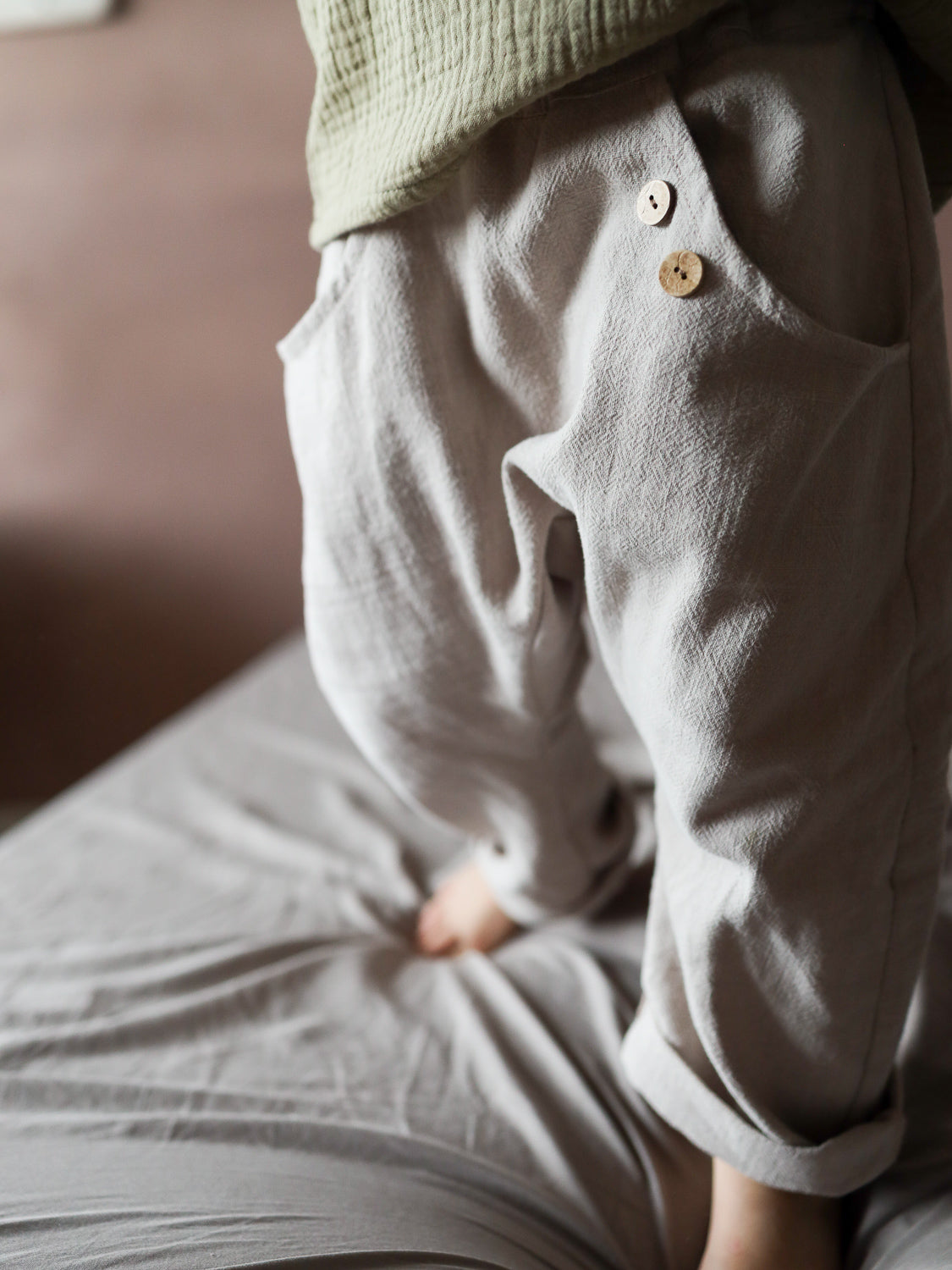 Kind steht barfuß auf Bett, trägt Sommerknicker Leinen - Sand. Leicht-beige, locker sitzende Hose mit Holzknöpfen. Kombiniert mit einem hellgrünen Oberteil. Perfekt für Bewegungsfreiheit und Komfort.