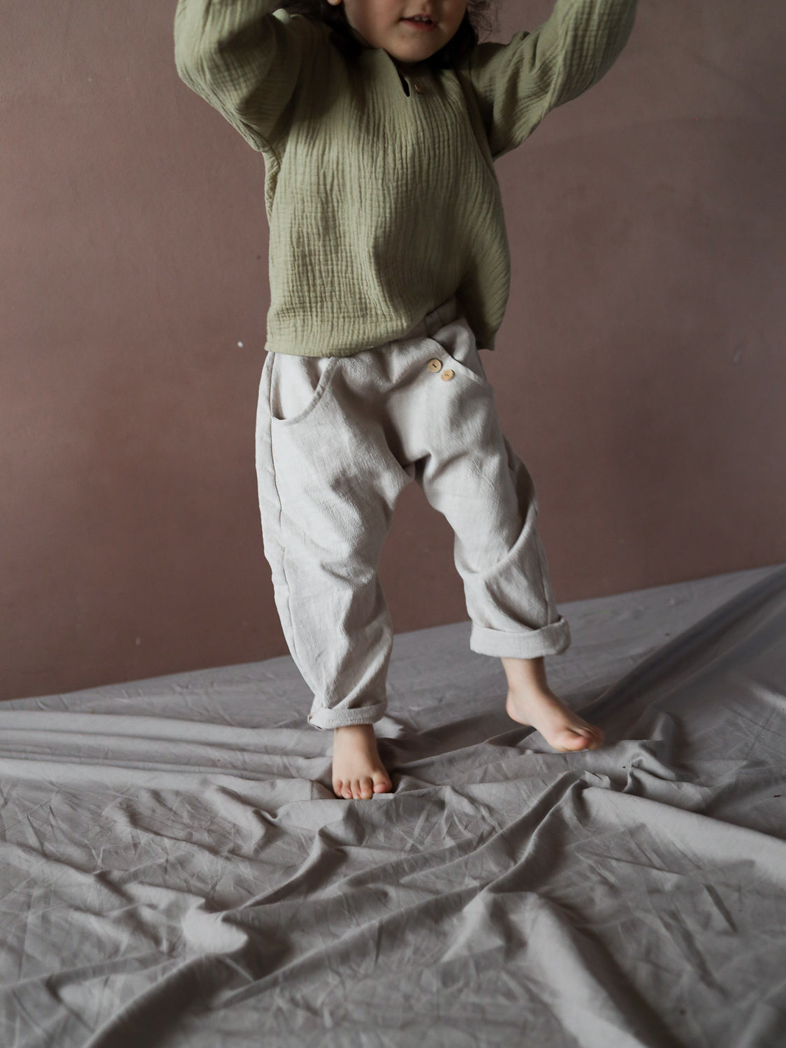 Kleinkind in olivegrünem Pullover und grauer Hose spielt auf grauem Stoff. Die Sommerknicker Leinen - Sand sind bequem, mit Knopfdetails und lockerer Passform für Bewegungsfreiheit.