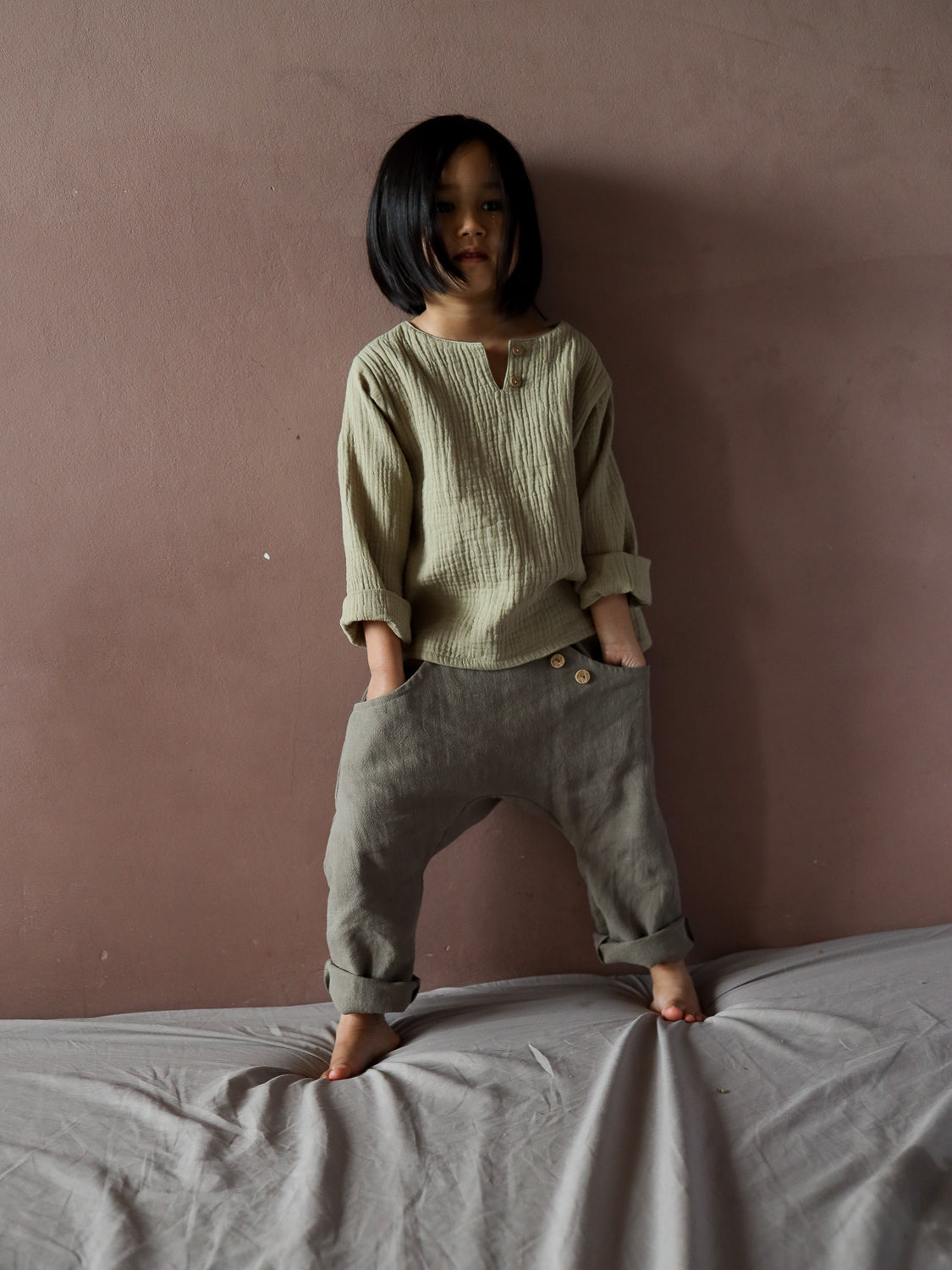 Kind in Sommerknicker Leinen - Oliv, locker geschnittene, olivfarbene Leinenhose mit umgekrempelten Bündchen, kombiniert mit beigem Langarmhemd. Steht barfuß auf grauem Untergrund vor einer rosafarbenen Wand.