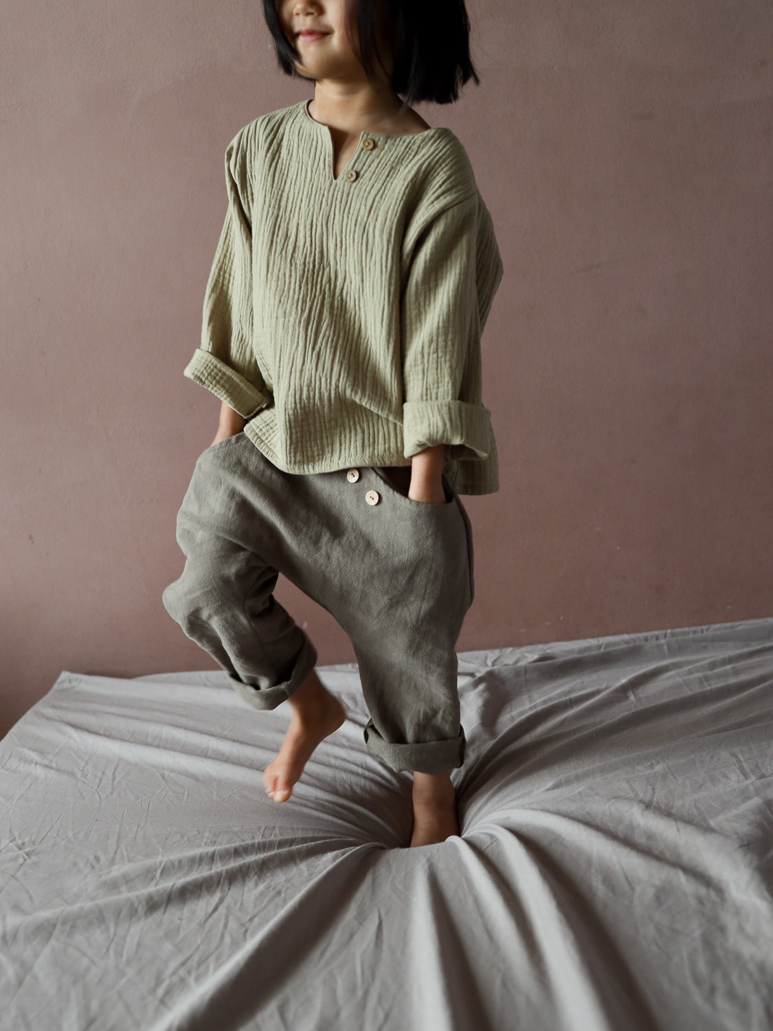 Kind springt auf Bett, trägt Sommerknicker Leinen - Oliv mit lockerem Schnitt, Knöpfen an der Tasche und Gummibund für Bewegungsfreiheit; perfekt für aktive Sommertage.