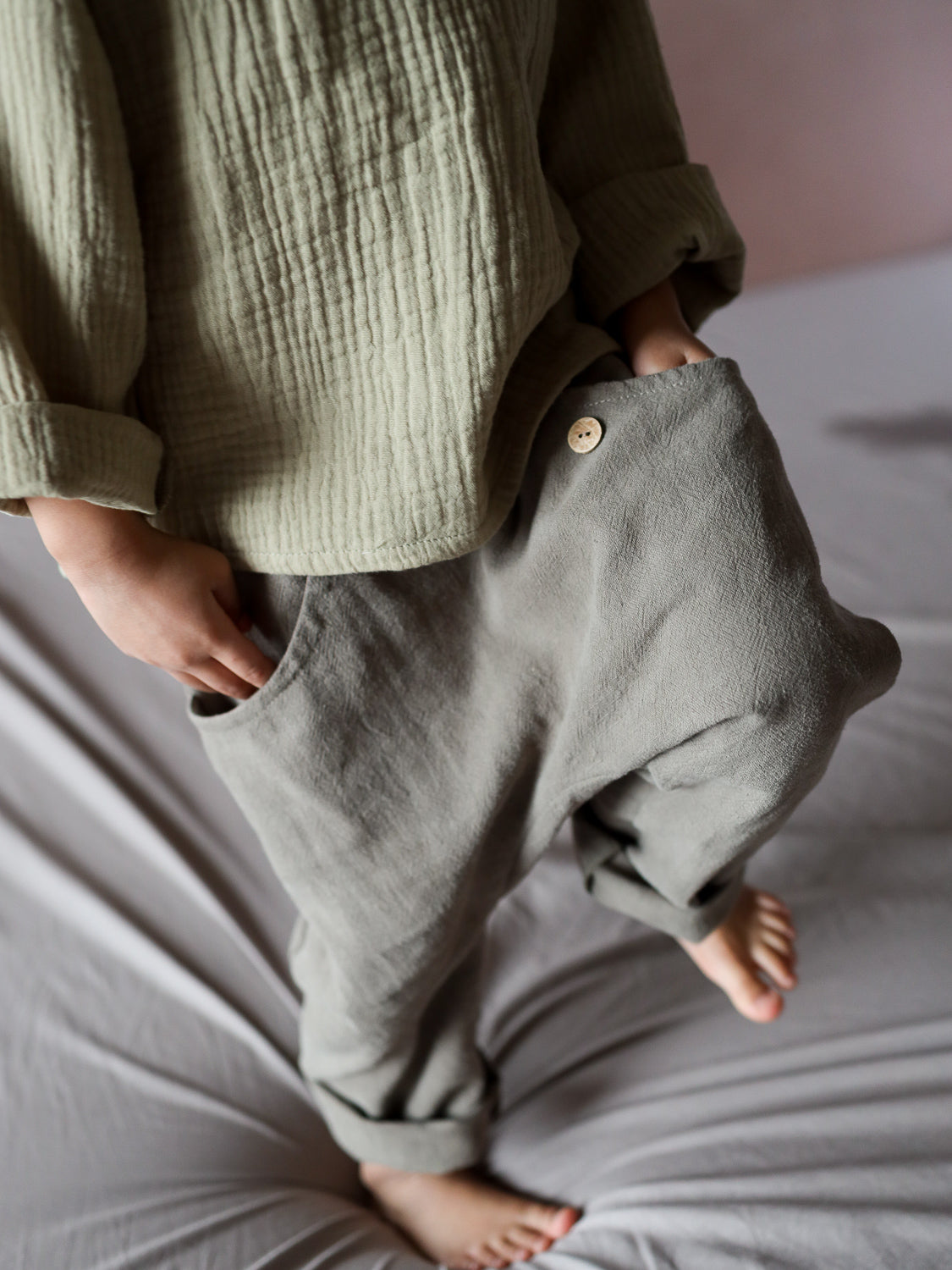 Kleinkind trägt Sommerknicker Leinen - Oliv: locker sitzende, olivfarbene Leinenhose mit Holzknopf und Gummibund, kombiniert mit langärmligem Baumwoll-Gauze-Top. Ideal für Bewegungsfreiheit und Komfort.