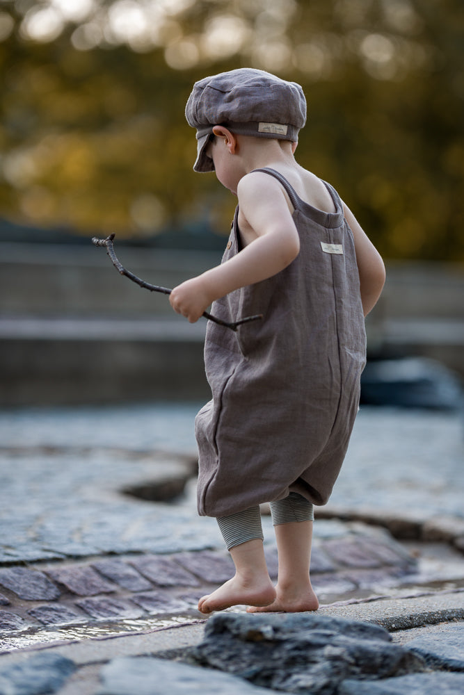 Ein Kind trägt die Schlupflatzhose Leinen - Oliv, steht barfuß auf nassem Stein, hält einen kleinen Stock und betrachtet Wasser. Die Hose ist locker, ohne Knöpfe, ideal für Entdecker.