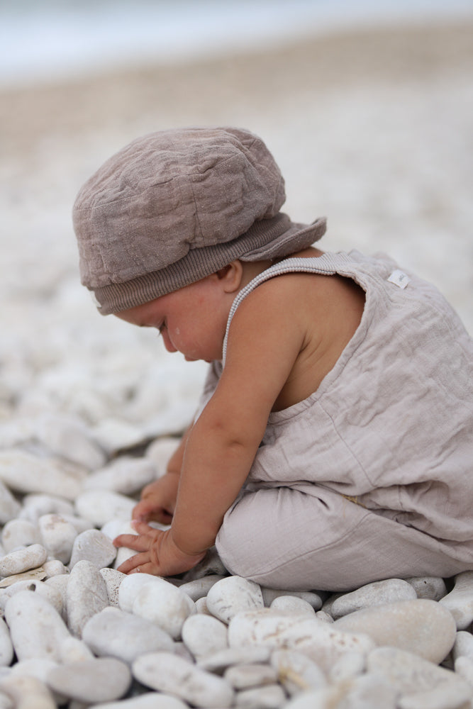 Ein Kind trägt die Kurze Schlupflatzhose Bio-Musselin - Sand, sitzt auf weißen Kieseln und untersucht neugierig die Steine mit den Händen. Der bequeme Einteiler eignet sich perfekt für sommerliche Abenteuer.