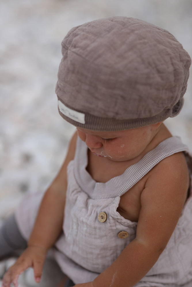 Kleinkind im sandfarbenen Kurze Schlupflatzhose Bio-Musselin, mit Holzknöpfen und passender Mütze. Es sitzt und betrachtet seine Hände, während leichte Flecken um Mund und Arme sichtbar sind.