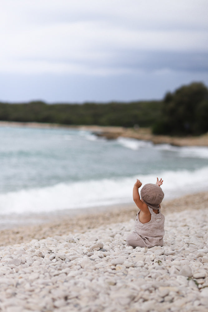 Kleinkind am Strand mit erhobenen Armen, trägt die Kurze Schlupflatzhose Bio-Musselin - Sand, eine bequeme, handgenähte Sommer-Einteilerhose aus Berlin, ideal für spielerische Bewegungsfreiheit.