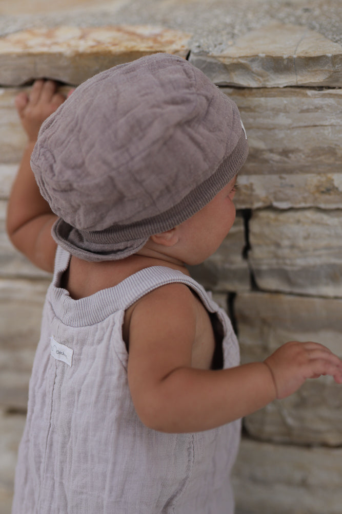 Ein Kleinkind trägt die Kurze Schlupflatzhose Bio-Musselin - Sand, steht an einer Steinmauer, stützt sich mit einer Hand ab. Der bequeme Einteiler ist ärmellos und ideal für Sommerabenteuer.