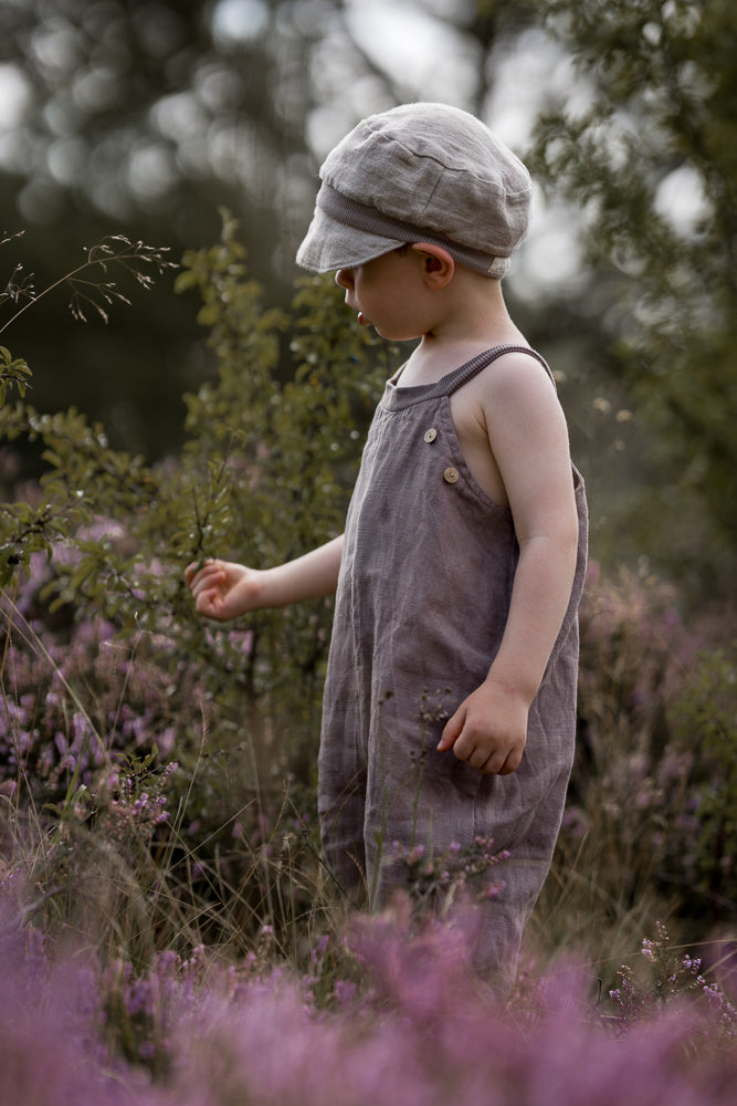 Kleinkind in einem Feld, das eine Schlupflatzhose Leinen - Oliv trägt, berührt sanft Pflanzen. Die ärmellose, locker sitzende Latzhose bietet viel Bewegungsfreiheit, ideal für Entdecker.