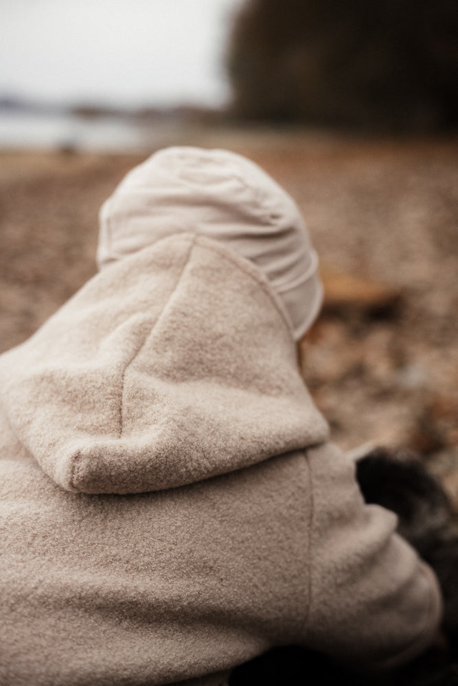Eine Person am Ufer trägt die Wichteljacke Wollwalk - Sand. Die Jacke ist aus Wollwalk gefertigt, mit Kapuze und Bio-Jersey-Futter, ideal für Herbst- und Wintermonate.