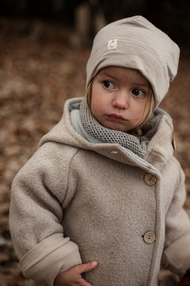 Kleinkind in der Wichteljacke Wollwalk - Sand mit großen Holzknöpfen und krempelbaren Bündchen, kombiniert mit einem grauen Schal und beiger Mütze, steht auf Herbstlaub.