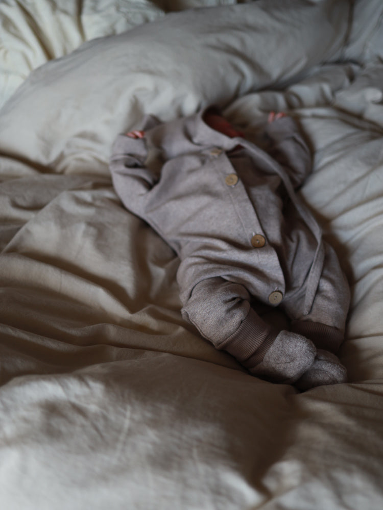 Ein Neugeborenes in einem Mini Overall Feinstrick - Hafer Meliert liegt auf einem neutralen Bett, trägt einen langärmligen, knöpfbaren Overall mit passenden Füßlingen, ideal für empfindliche Babyhaut.
