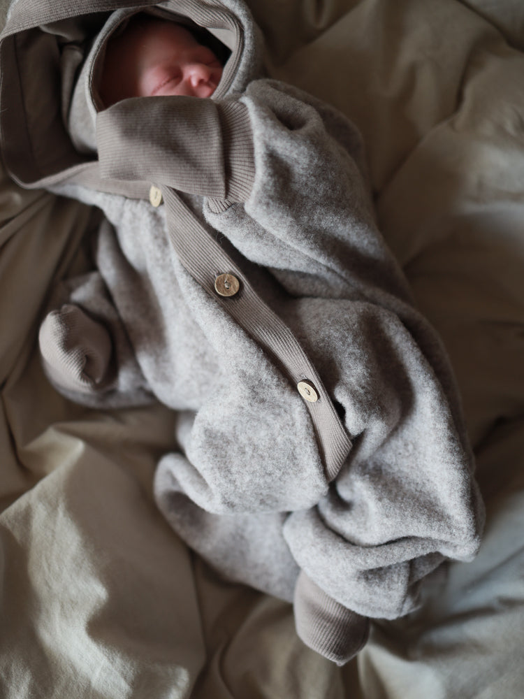 Kuschelanzug Merino Wollfleece - Hafer: Ein Baby schläft in einem weichen, heathered grauen Einteiler mit Knopfleiste und gerippten Bündchen, auf einer neutralen Decke. Perfekt für Komfort und Schutz.