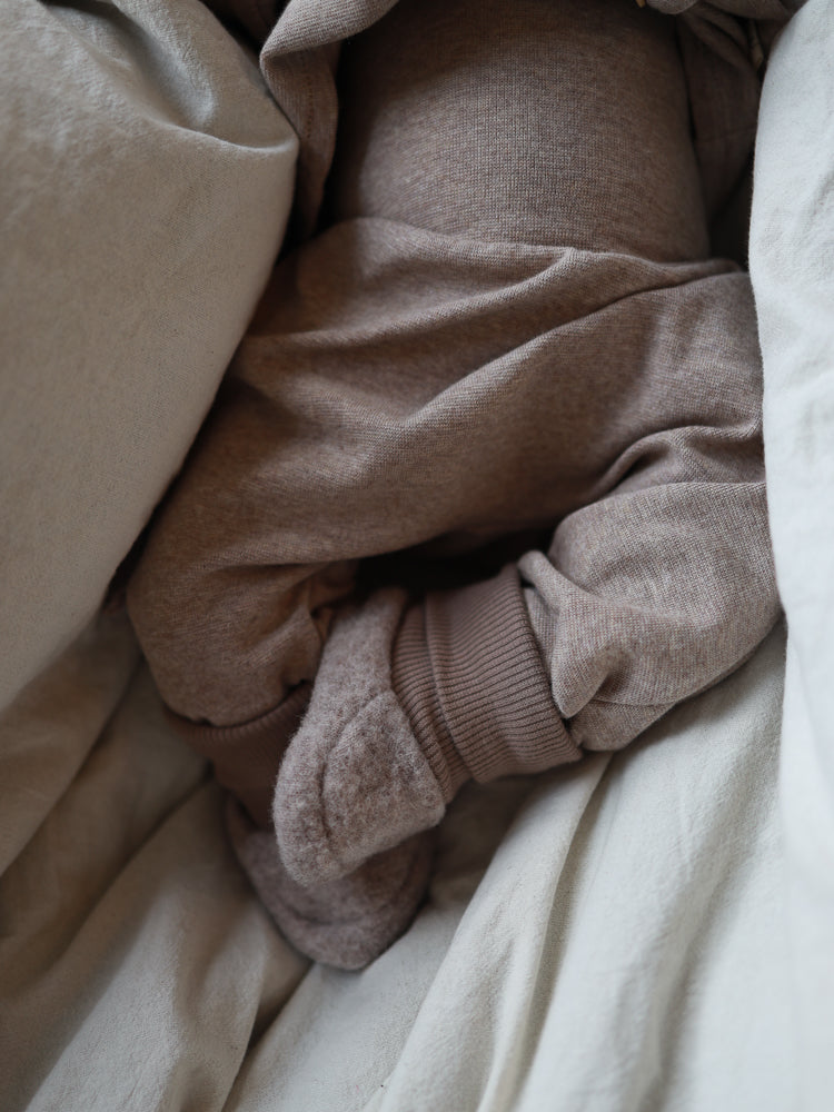Babybooties Merino Wollfleece - Hafer: Ein Baby trägt gemütliche Merino-Wollfleece-Booties, gebettet in weichen Decken. Die Booties wirken flexibel und halten die Füße warm, passend zu jedem Outfit.