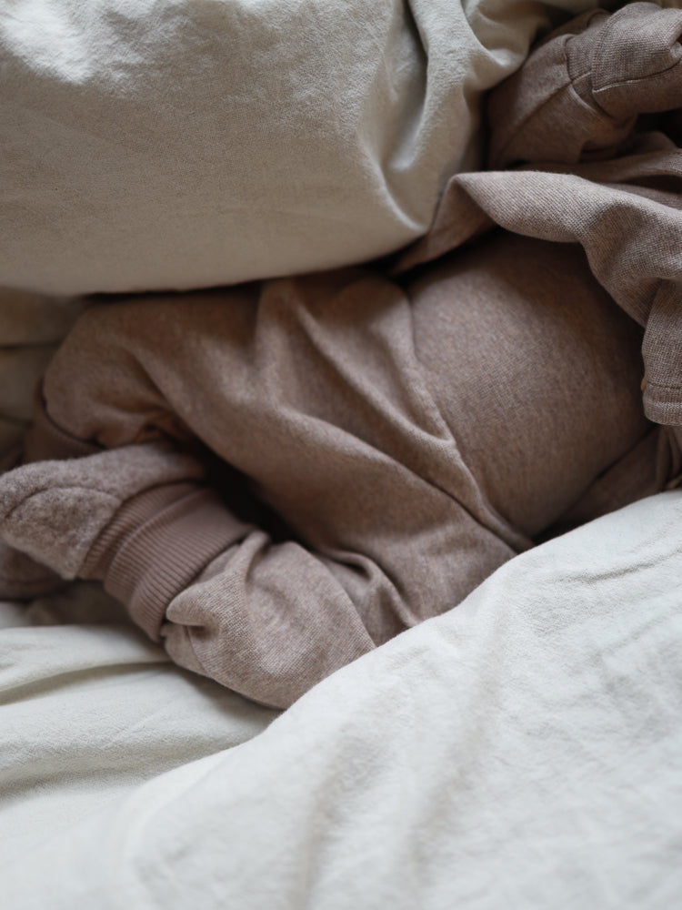 Ein Baby trägt Babyknicker Feinstrick - Hafer Meliert, aus weichem, atmungsaktivem Feinstrick-Jersey. Die melierte Hose sitzt locker und bequem, ideal zum Kuscheln, mit cremigen Bettzeug umgeben.