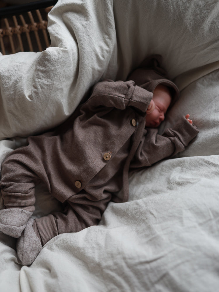 Neugeborenes in Mini Overall Feinstrick - Praline Meliert, mit Kapuze und Knöpfen, liegt auf weißem, weichem Bettzeug. Der Overall bietet Bewegungsfreiheit und ein angenehmes Tragegefühl.