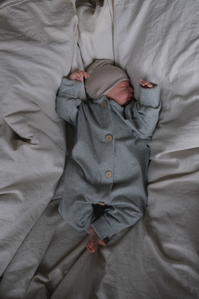 Neugeborenes schläft in einem Mini Overall Feinstrick - Mint Meliert, mit langen Ärmeln und Knopfleiste, auf weichem Bett, trägt auch eine blassgraue/beige Strickmütze.