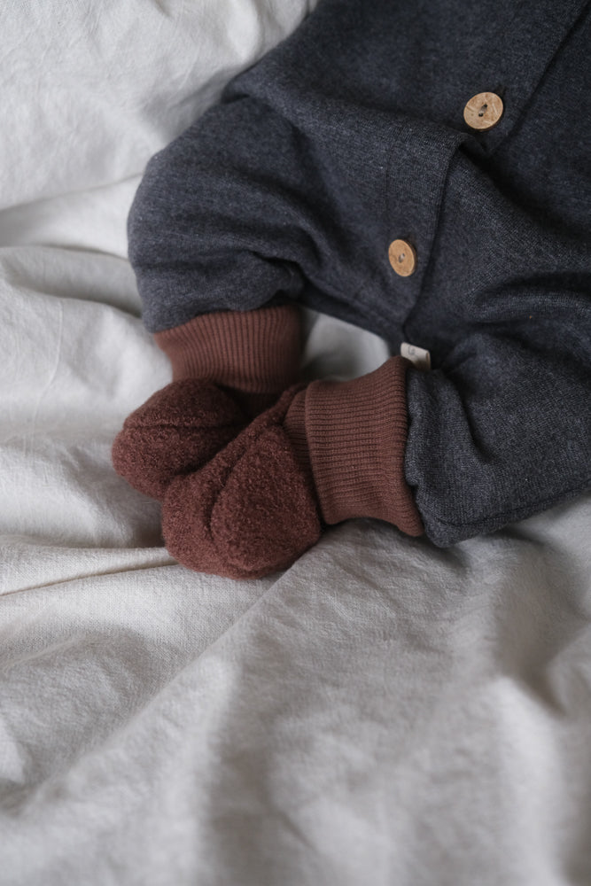 Nahaufnahme von Babybeinen in einem dunkelgrauen Einteiler mit Holzknöpfen. Sie tragen Babybooties Wollwalk - Schoko, aus gewalkter Schurwolle, handgenäht in Berlin, mit elastischen Bündchen für optimalen Halt.