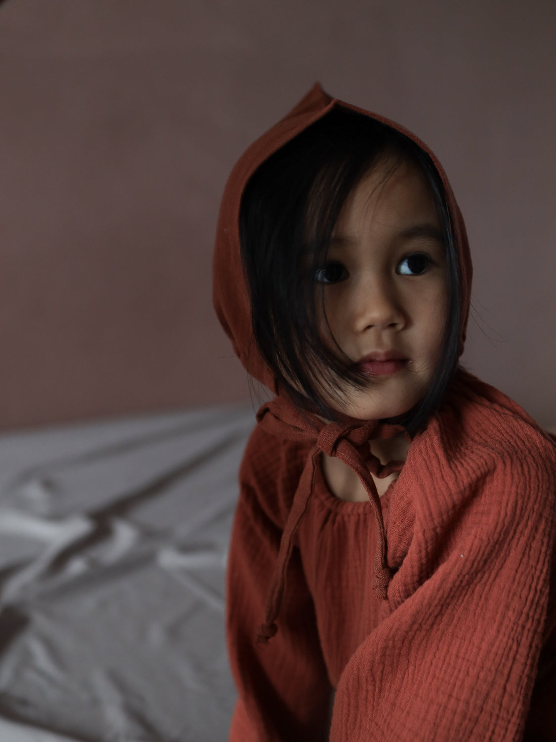 Ein Kleinkind sitzt auf einem Bett, trägt eine Zwergenmütze Leinen - Ziegel, die spitz zuläuft und Kopf und Ohren bedeckt, ergänzt durch einen passenden, weichen Overall.