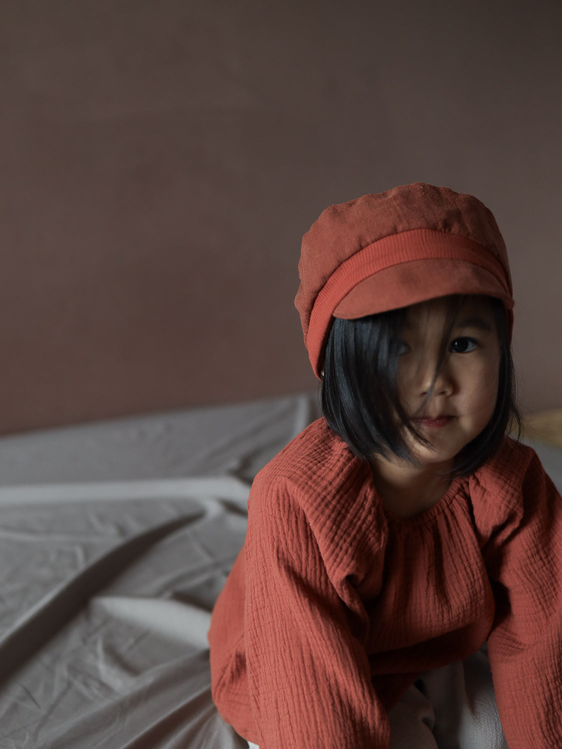 Ein Kleinkind trägt eine Michelmütze Leinen - Ziegel, die rustikal und strukturiert ist. Passend dazu trägt es ein langärmeliges Oberteil in ähnlichem Design, während es auf einem zerknitterten, hellen Laken sitzt.