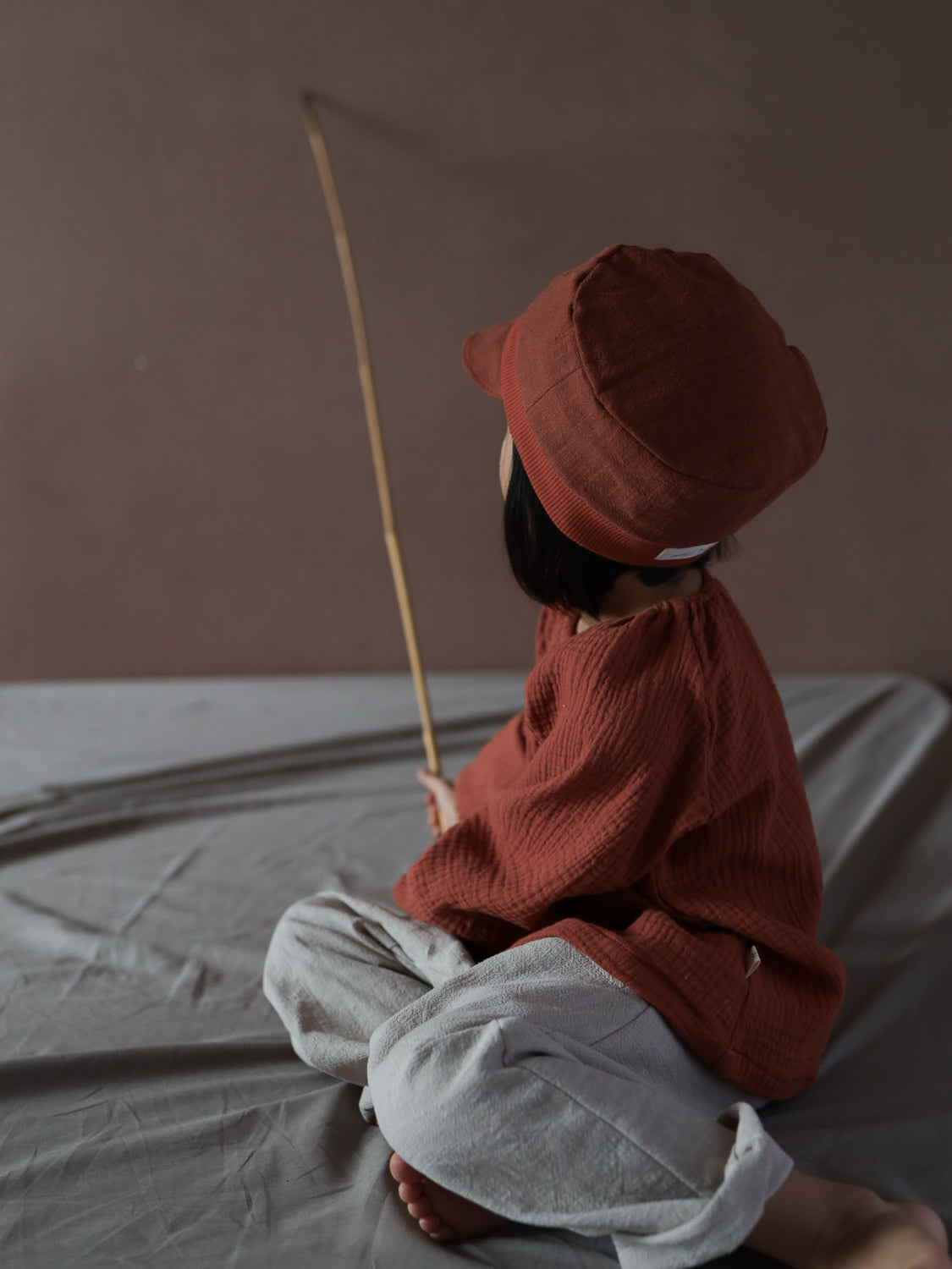 Ein Kind sitzt auf einer weichen Oberfläche, trägt die Michelmütze Leinen - Ziegel. Es hat ein strukturiertes, rostfarbenes Langarmshirt und eine passende Mütze, kombiniert mit locker sitzenden hellgrauen Hosen.