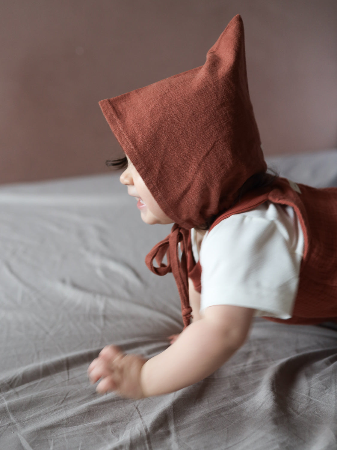 Ein Kleinkind krabbelt auf weicher Oberfläche, trägt die Zwergenmütze Leinen - Ziegel. Die spitz zulaufende Mütze schützt Kopf und Ohren, kombiniert mit passenden Overalls und weißem Shirt.