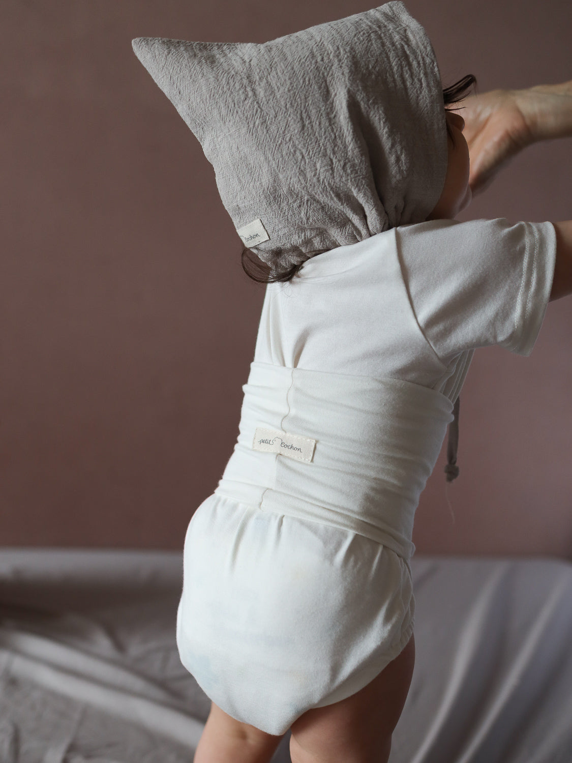 Ein Kleinkind trägt die Zwergenmütze Leinen - Sand, die Kopf und Ohren schützt. Der weiche, dehnbare Stoff sorgt für Komfort, während das Kind sich auf einer gepolsterten Oberfläche abstützt.