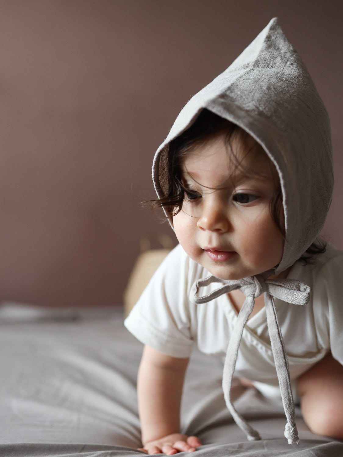 Ein Baby in einem hellen Strampler trägt eine Zwergenmütze Leinen - Sand, die aus weichem, atmungsaktivem Feinstrick-Jersey besteht. Die Mütze schützt Kopf und Ohren beim Krabbeln.